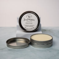 An open tin of cream colored balm next to a tin lid with a label that says Lakota Made Just Wakan cologne balm. The tins are on a gray marble surface in front of a white background. The ingredients are shea butter, beeswax, sunflower oil, patchouli and cedarwood essential oil. 