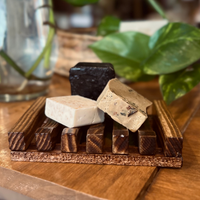 A brown bamboo soap dish with three small soap samples in front of a glass vase and green leaves