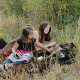 Two girls petting a brown dog in a grass and flower field