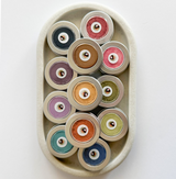 A white stone oval dish with twelve round aluminum tins of brightly colored eyeshadows. The labels read Lakota Made War Paint Makeup.