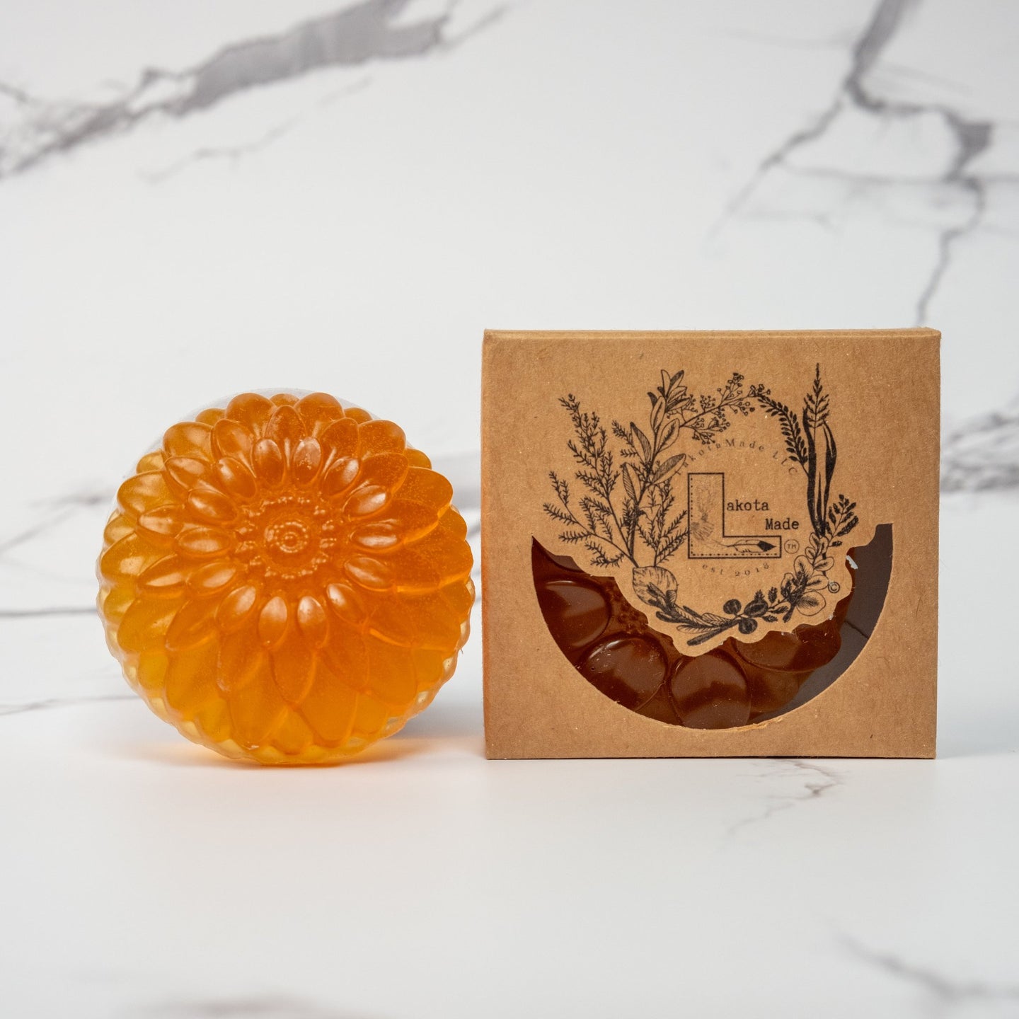 A round flower shaped translucent orange colored soap bar with a second bar in a brown cardboard package. The package says Lakota Made. The products are on a white marble background.