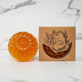 A round flower shaped translucent orange colored soap bar with a second bar in a brown cardboard package. The package says Lakota Made. The products are on a white marble background.