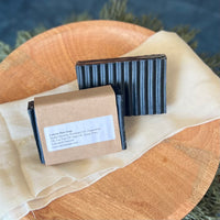 A rectangular grooved black colored soap bar with a second bar in a brown paper sleeve. The package says Lakota Made Lakota Man Soap. The products are on a cloth in a wooden dish surrounded by pine needles.