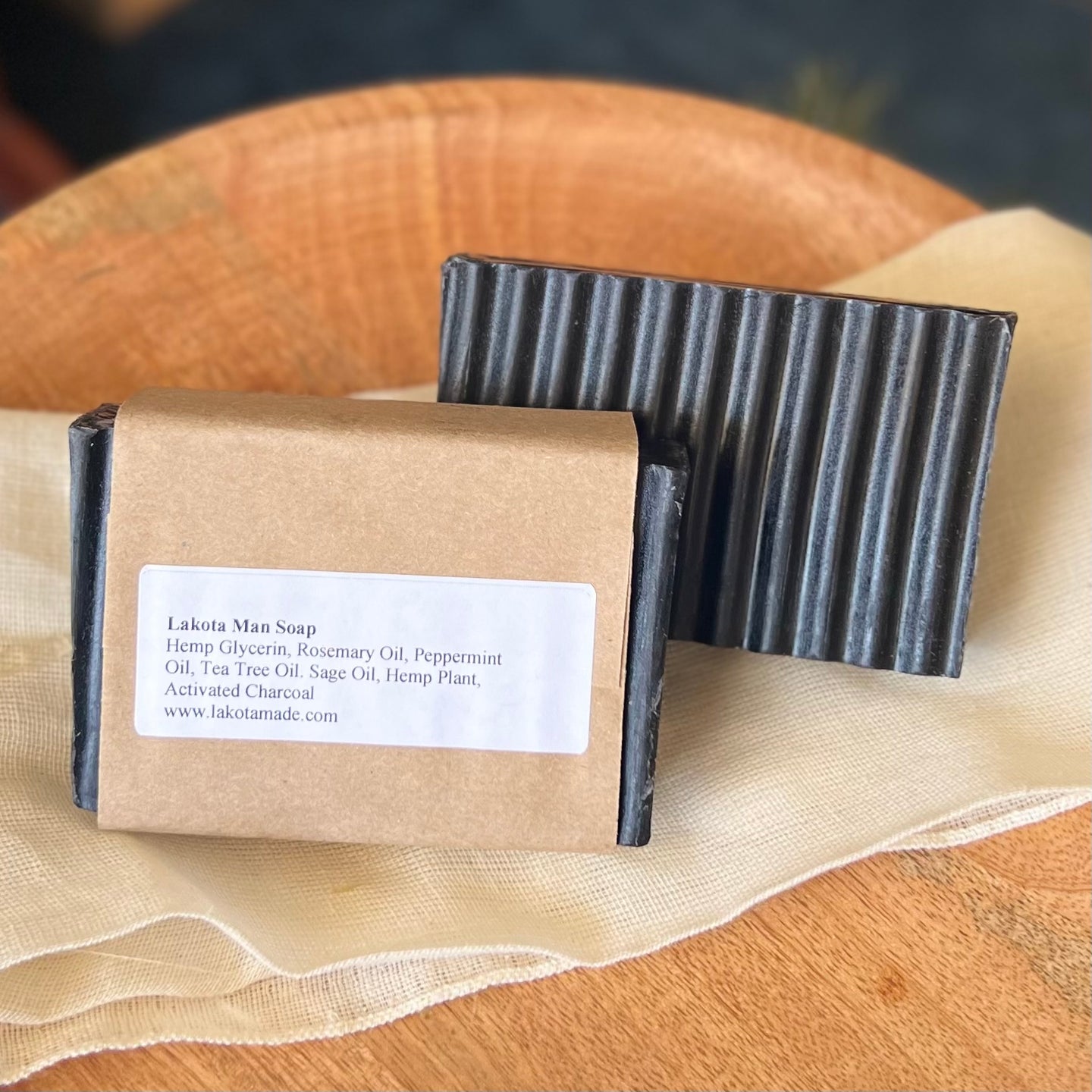 A rectangular grooved black colored soap bar with a second bar in a brown paper sleeve. The package says Lakota Made Lakota Man Soap. The products are on a cloth in a wooden dish surrounded by pine needles.