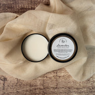 A round black aluminum tin containing a cream colored solid lotion. The lid has a round white label that says Lavender Luxury Solid Lotion ingredients Shea Butter, Beeswax, Lavender Essential Oil. The tin and lid are on a muslin fabric on top of a rustic wood surface.