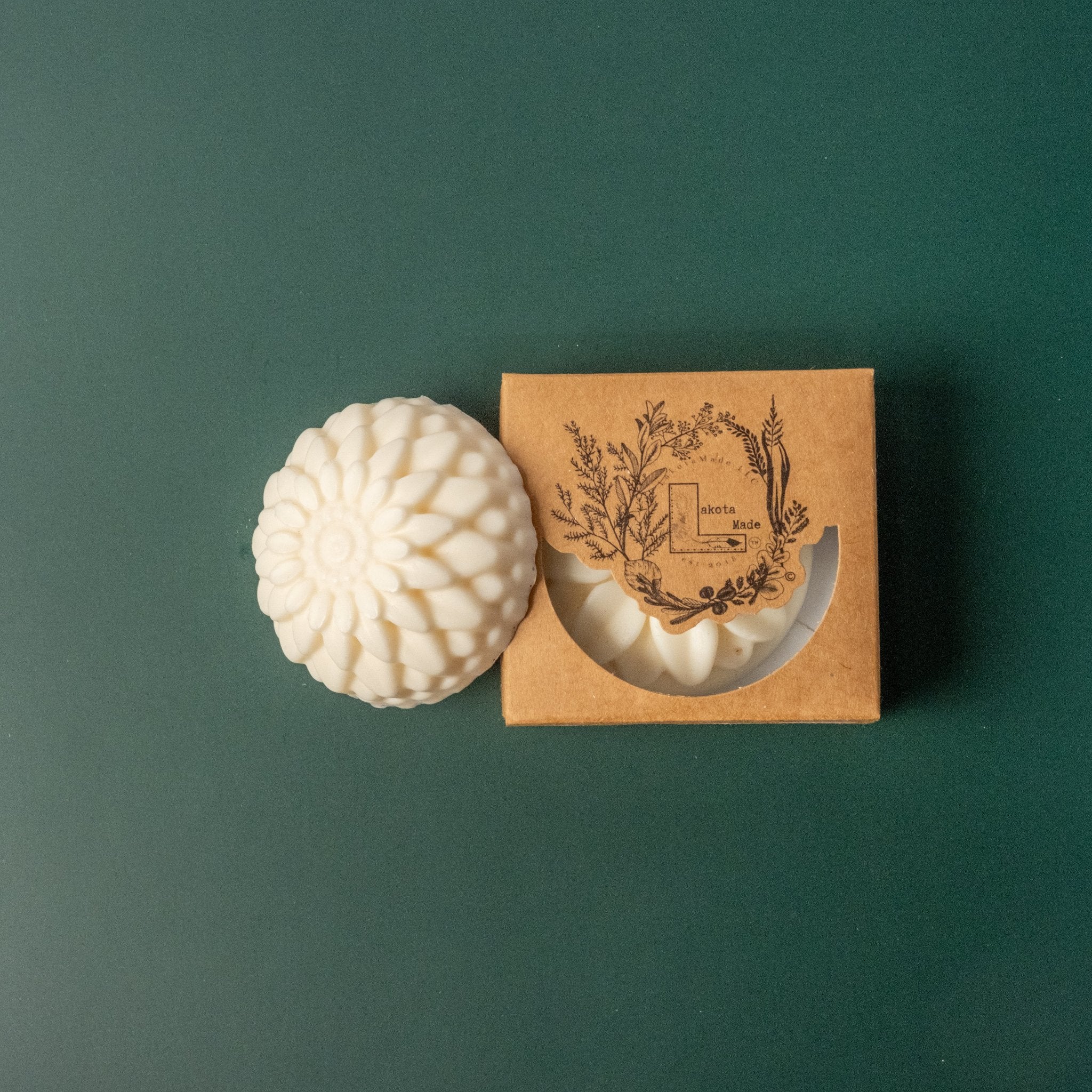 A round flower shaped white soap bar with a second bar in a brown cardboard package. The package says Lakota Made. The products are on a dark green background.