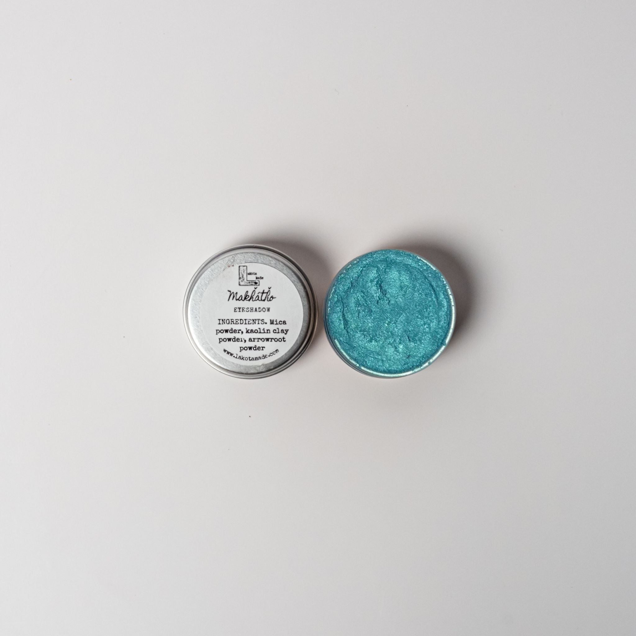 An open aluminum tin of vibrant light blue powder on a white background with a white label that says Lakota Made Makhatho Blue Earth Eyeshadow Ingredients mica powder, kaolin clay powder, arrowroot powder.