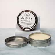 An open tin of cream colored balm next to a tin lid with a label that says Lakota Made Makhu Rub balm. The tins are on a gray marble surface in front of a white background. The ingredients are shea butter, beeswax, sunflower oil, menthol crystals, eucalyptus and peppermint essential oil.