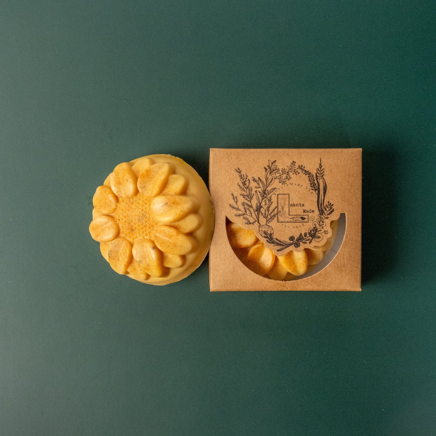 A round flower shaped yellow soap bar with a second bar in a brown cardboard package. The package says Lakota Made. The products are on a dark green background.