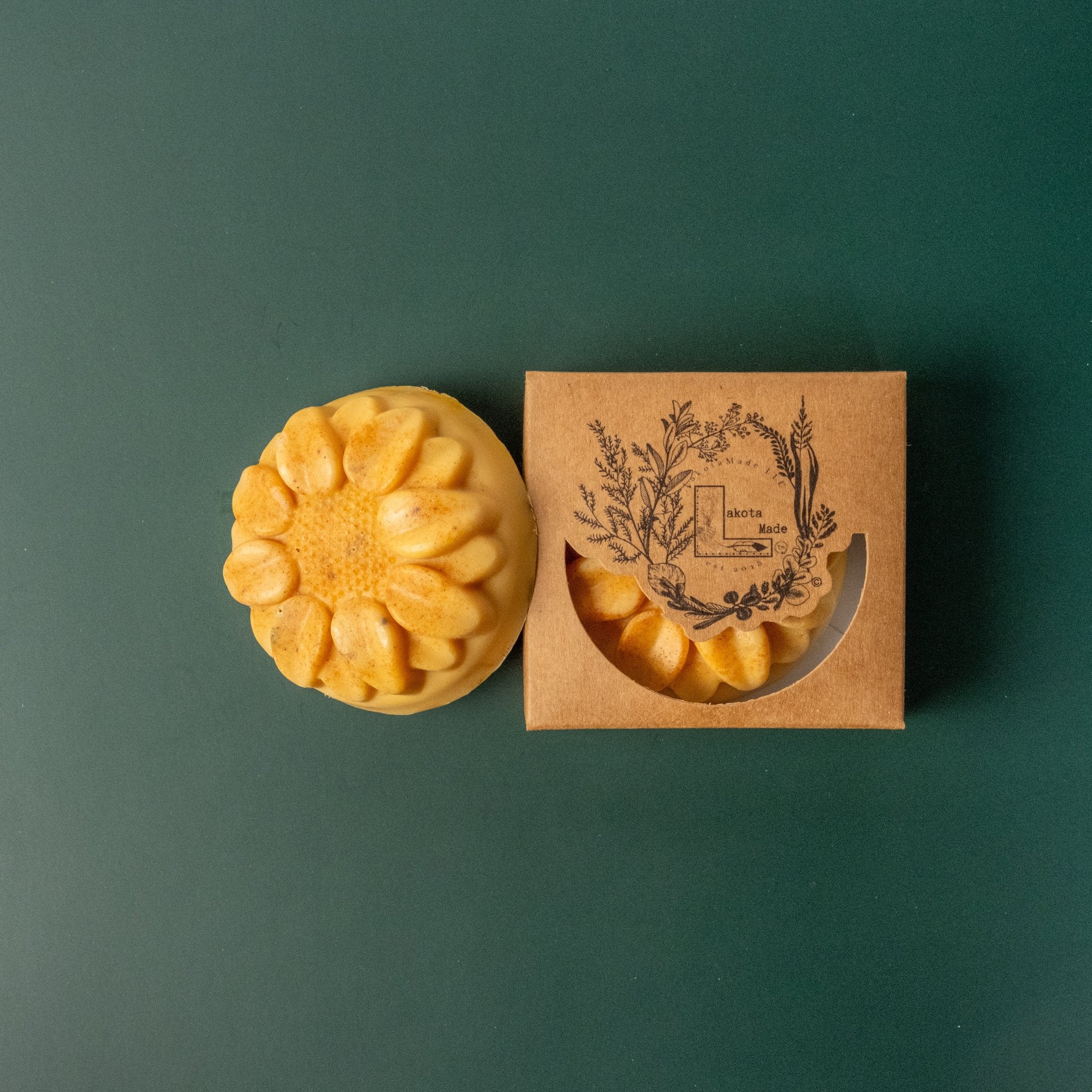 A round flower shaped yellow soap bar with a second bar in a brown cardboard package. The package says Lakota Made. The products are on a dark green background.