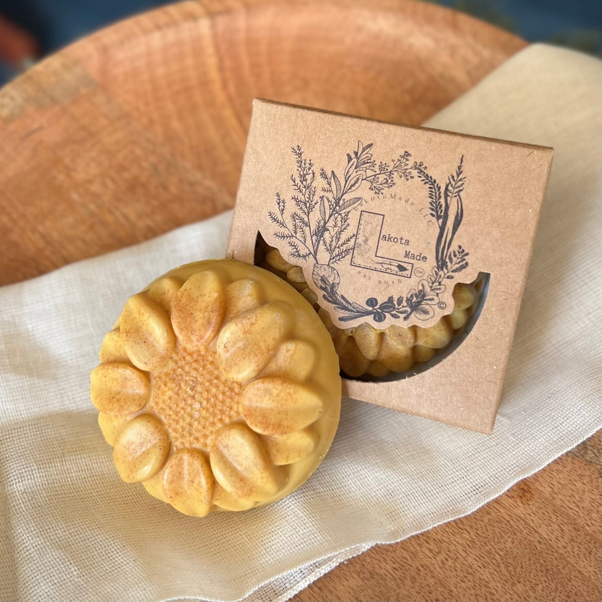 A close up of a round flower shaped yellow speckled colored soap bar with a second bar in a brown cardboard package. The package says Lakota Made. The products are on a cloth in a wooden dish surrounded by pine needles.