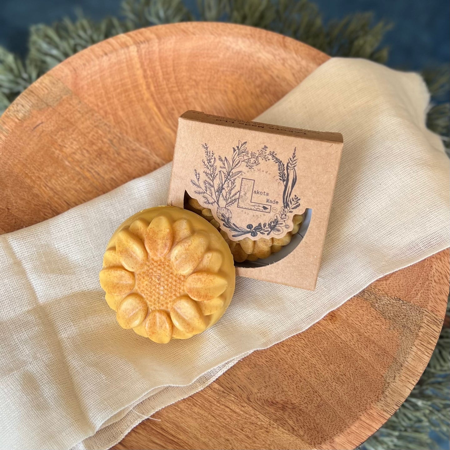 A round flower shaped yellow speckled colored soap bar with a second bar in a brown cardboard package. The package says Lakota Made. The products are on a cloth in a wooden dish surrounded by pine needles.