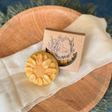 A round flower shaped yellow speckled colored soap bar with a second bar in a brown cardboard package. The package says Lakota Made. The products are on a cloth in a wooden dish surrounded by pine needles.