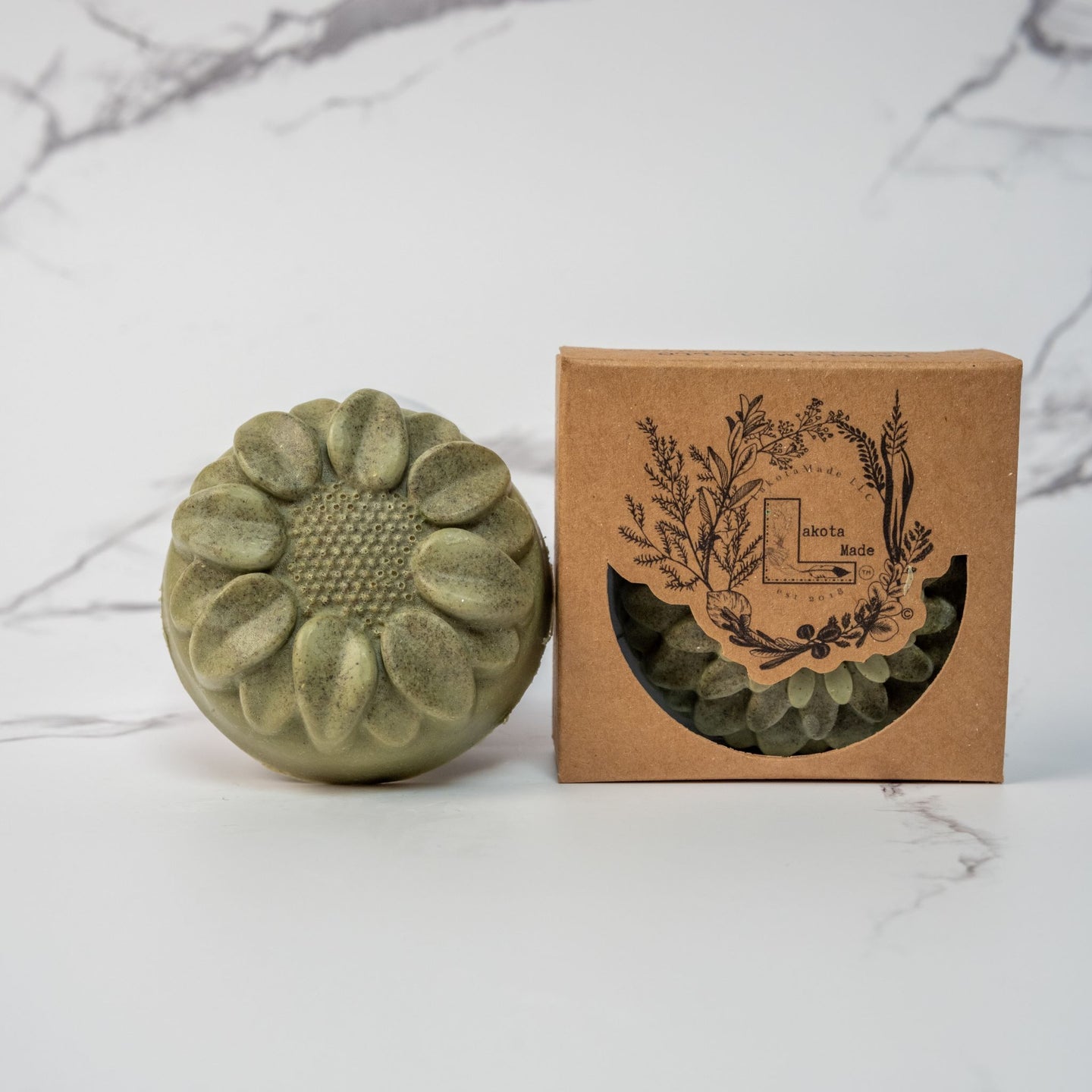 A round flower shaped sage green colored soap bar with a second bar in a brown cardboard package. The package says Lakota Made. The products are on a white marble background.