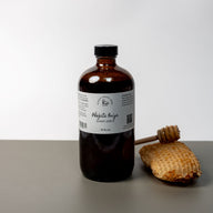 A large brown bottle with a white label that says Lakota Made Phezuta Oniya Honey Syrup 16 fl oz. Ingredients are raw honey, elderberries, mullein leaves, white willow bark. There is a honey comb and dipper next to the bottle. It is resting on a gray table top in front of a white background.