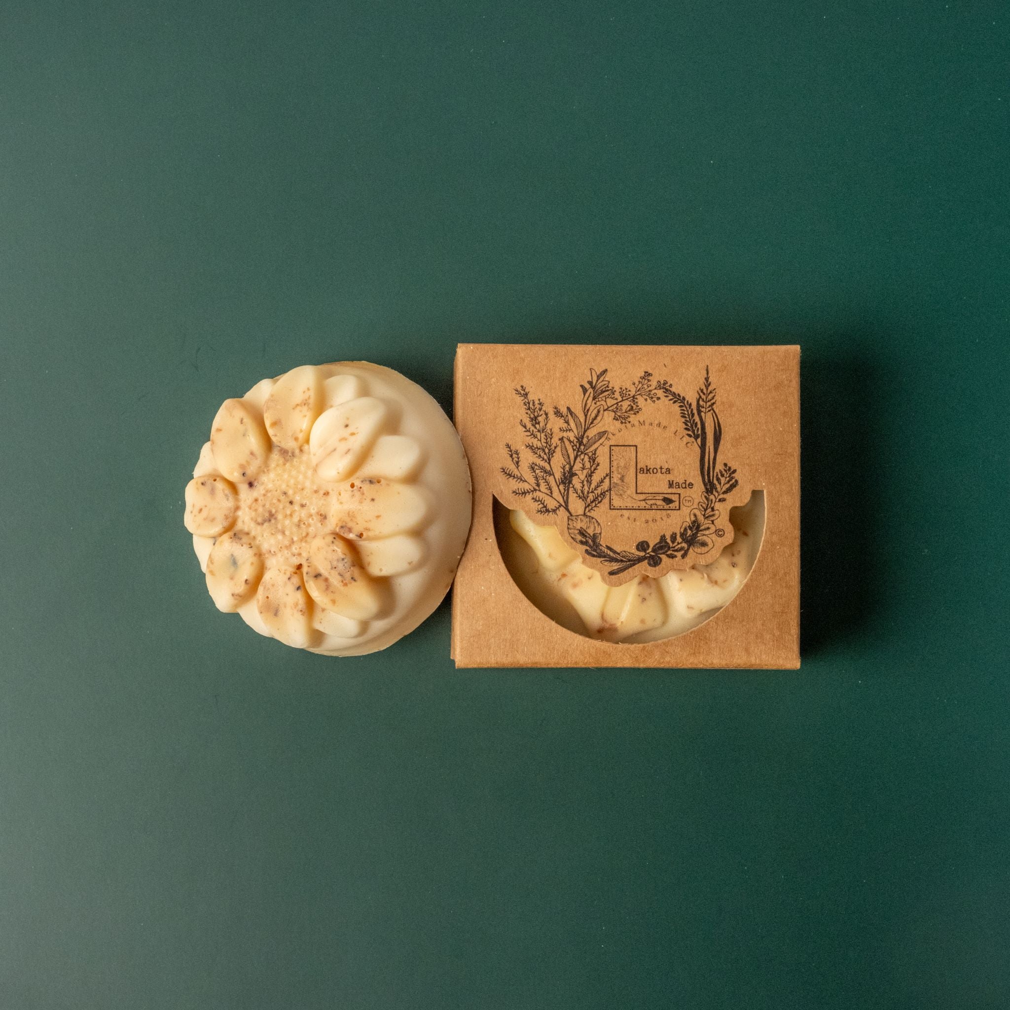 A round flower shaped cream and brown speckled soap bar with a second bar in a brown cardboard package. The package says Lakota Made. The products are on a dark green background.