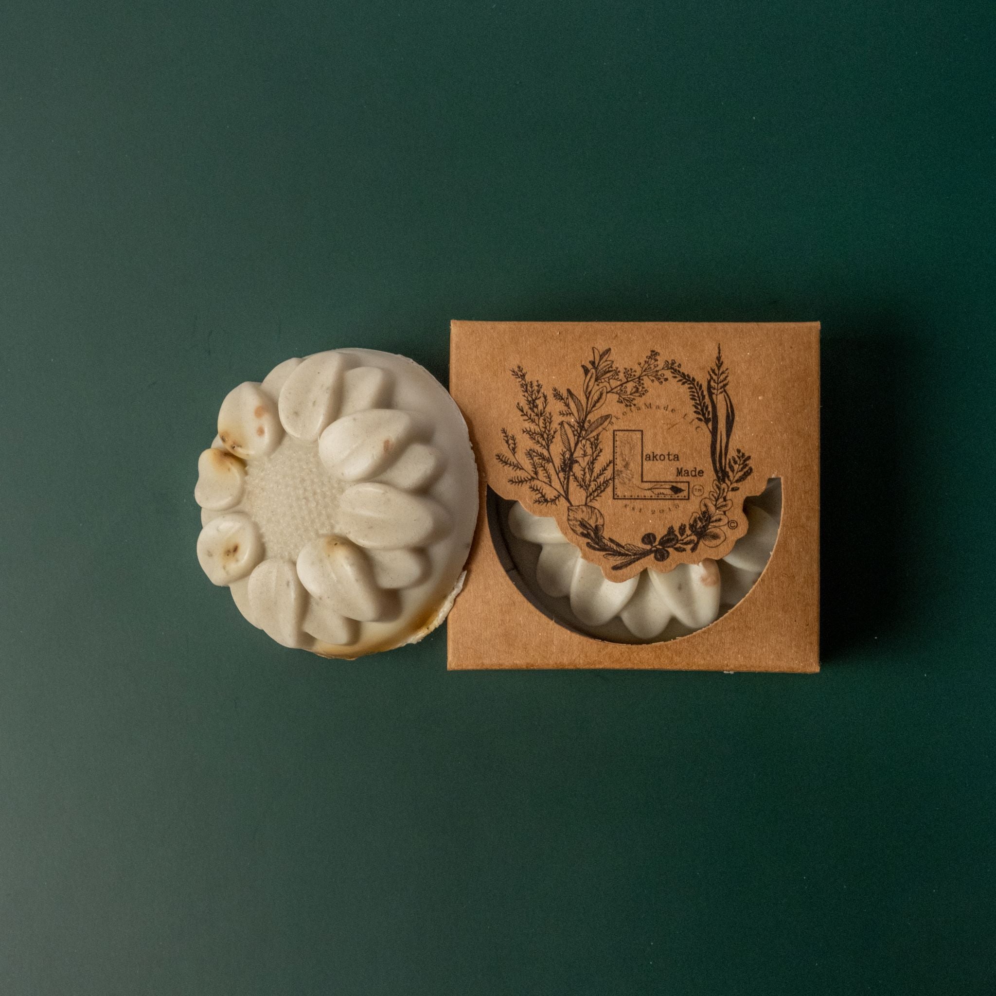 A round flower shaped gray and brown spotted soap bar with a second bar in a brown cardboard package. The package says Lakota Made. The products are on a dark green background.