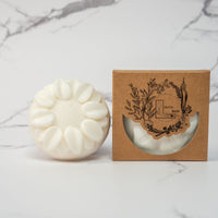 A round flower shaped cream colored soap bar with a second bar in a brown cardboard package. The package says Lakota Made. The products are on a white marble background.