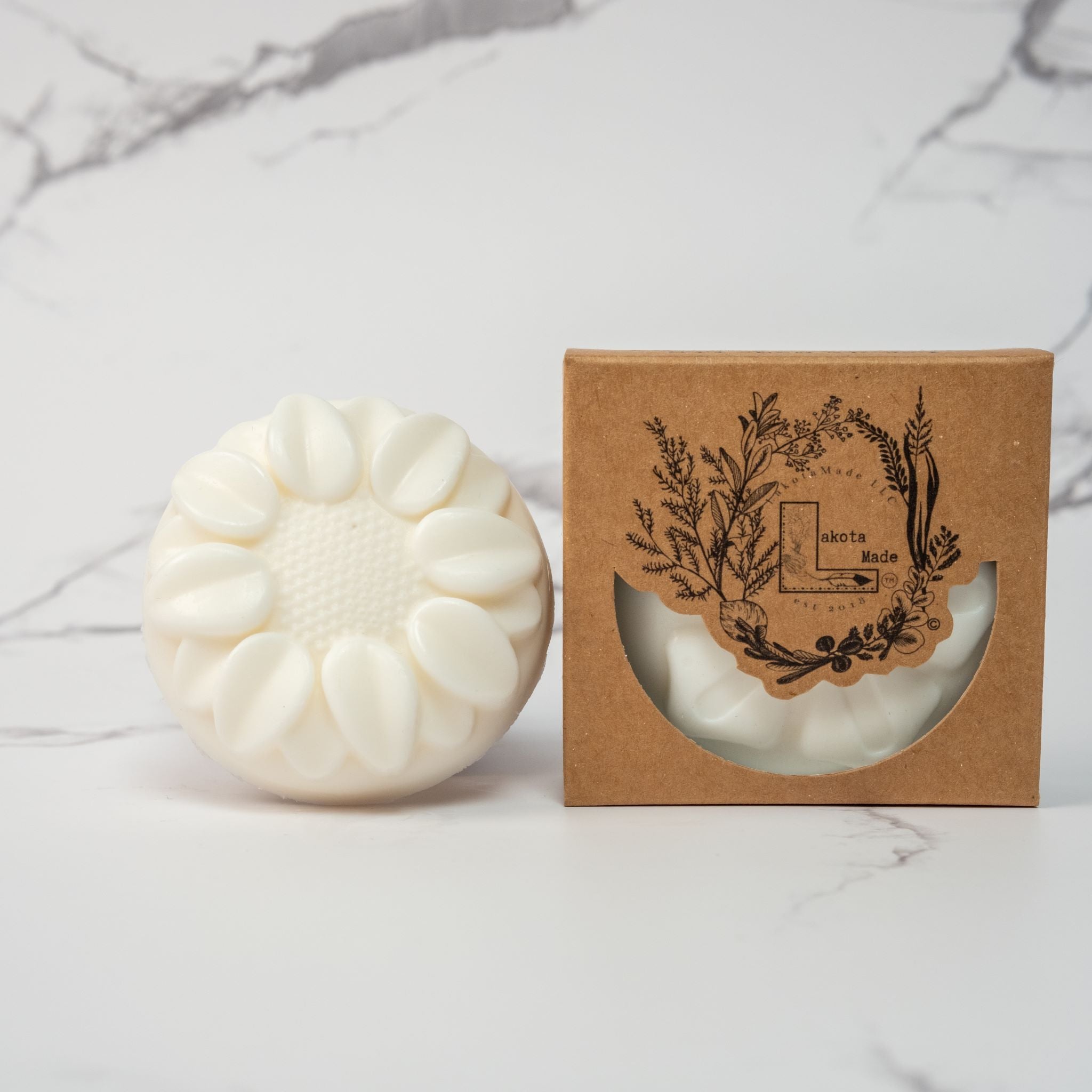 A round flower shaped cream colored soap bar with a second bar in a brown cardboard package. The package says Lakota Made. The products are on a white marble background.