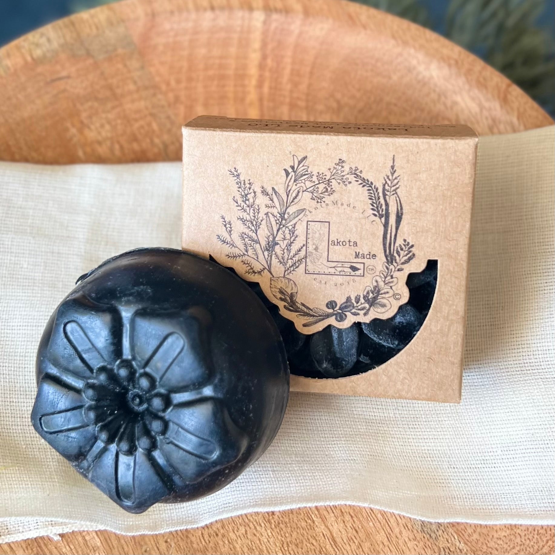 A close up of a round flower shaped black colored soap bar with a second bar in a brown cardboard package. The package says Lakota Made. The products are on a cloth in a wooden dish surrounded by pine needles.