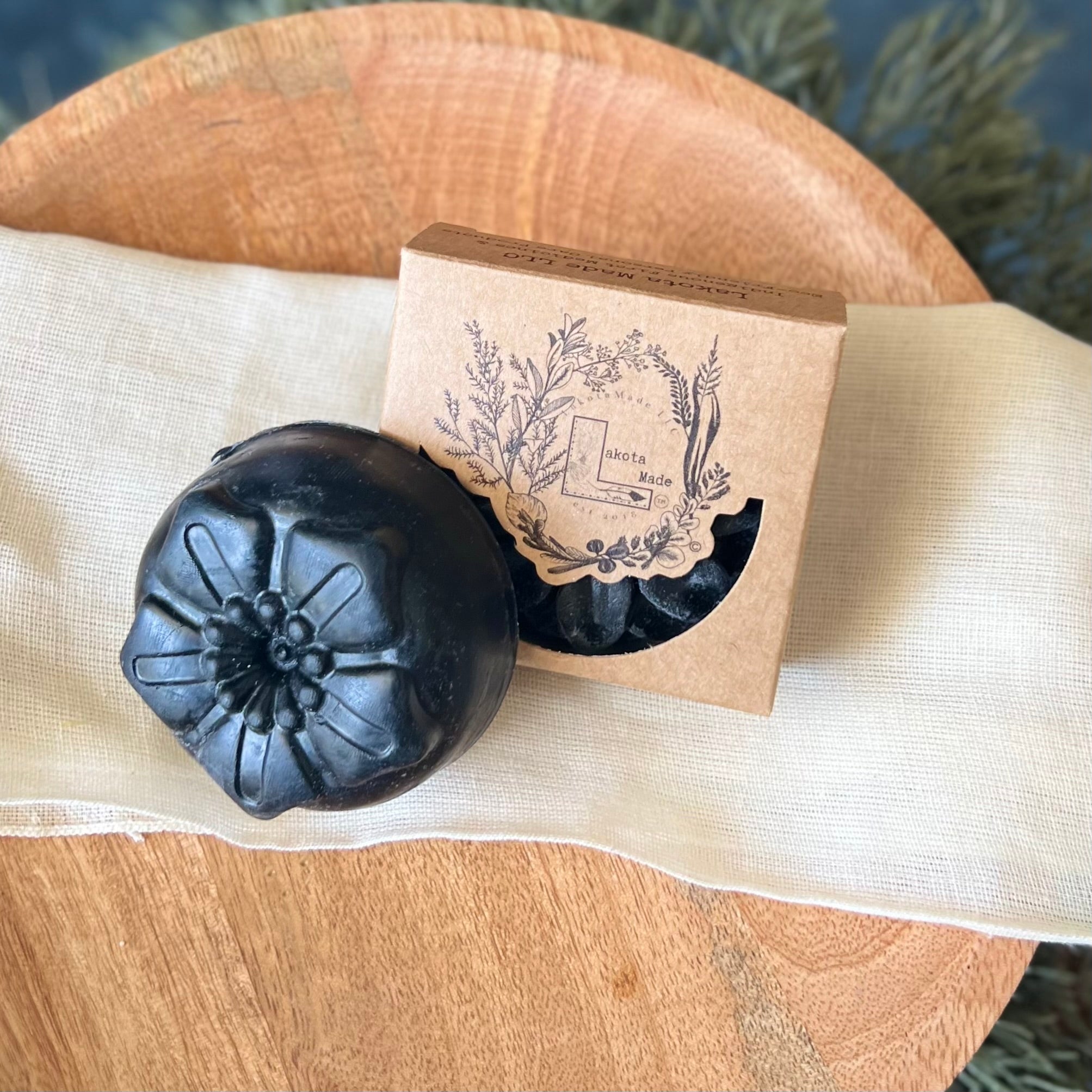 A round flower shaped black colored soap bar with a second bar in a brown cardboard package. The package says Lakota Made. The products are on a cloth in a wooden dish surrounded by pine needles.