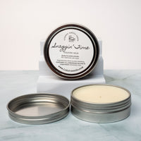 An open tin of cream colored balm next to a tin lid with a label that says Lakota Made Snaggin' Time cologne balm. The tins are on a gray marble surface in front of a white background. The ingredients are shea butter, beeswax, sunflower oil, cedarwood, tea tree, and peppermint essential oil. 