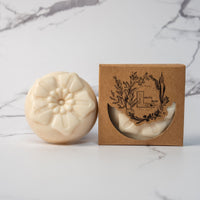 A round flower shaped cream colored soap bar with a second bar in a brown cardboard package. The package says Lakota Made. The products are on a white marble background.