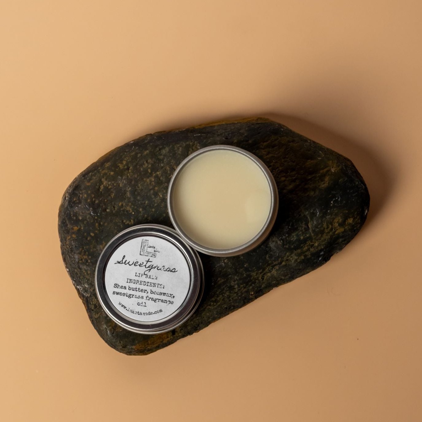 A round aluminum tin containing a cream colored lip balm. The tin has a lid with a round white label that says Lakota Made Sweetgrass Lip Balm ingredients Shea Butter, Beeswax, Sweetgrass Fragrance Oil. The tin and lid are positioned on a dark brown rock on a cream orange background.