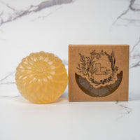 A round flower shaped translucent yellow colored soap bar with a second bar in a brown cardboard package. The package says Lakota Made. The products are on a white marble background.