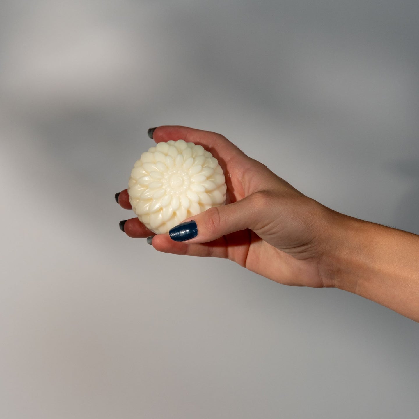 A woman's hand holding a round flower shaped cream colored soap bar on a gray background.