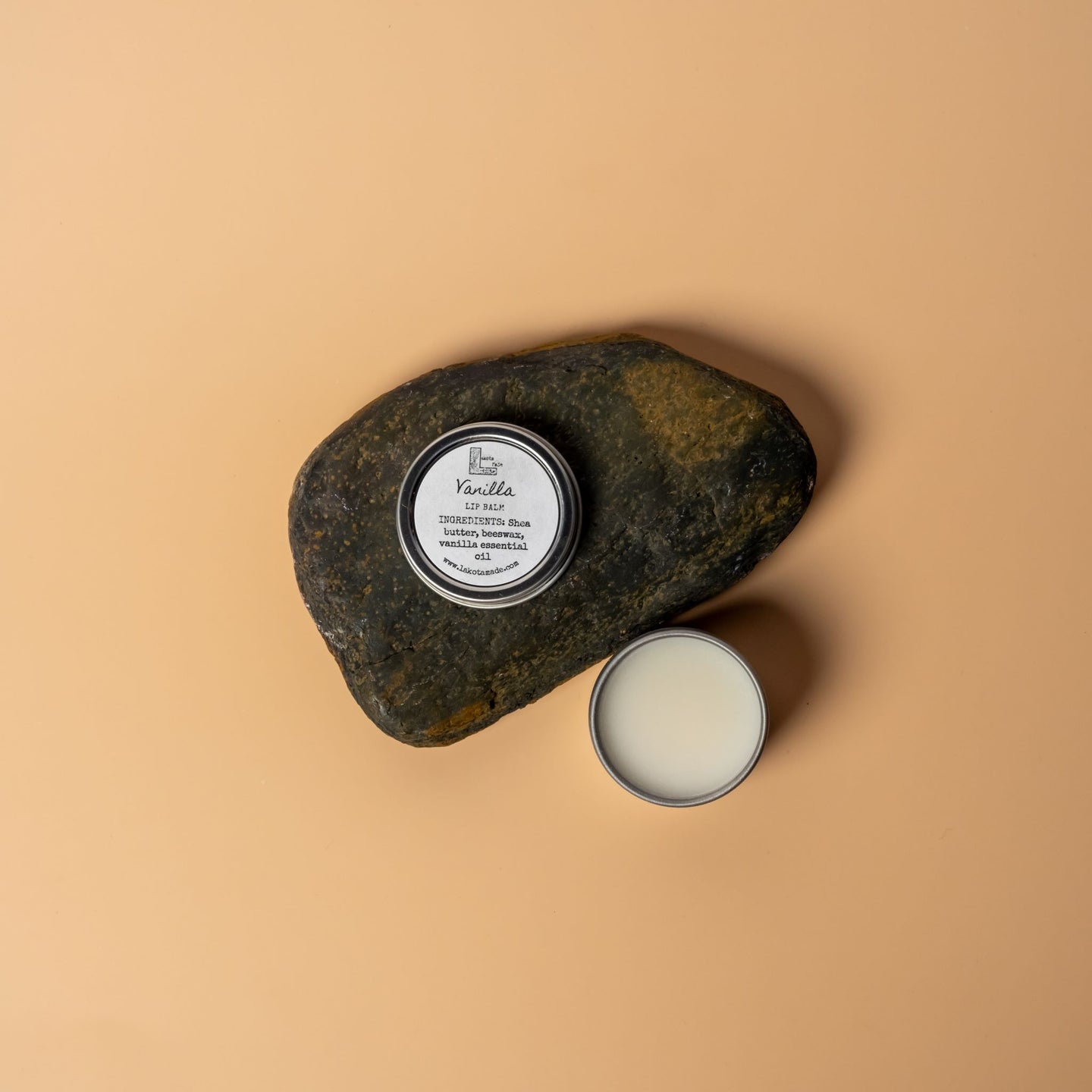 A round aluminum tin containing a cream colored lip balm. The lid says vanilla lip balm ingredients shea butter beeswax vanilla essential oil. The lid is positioned on a dark brown rock on a cream orange background.