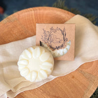 A round flower shaped cream colored conditioner soap bar with a second bar in a brown cardboard package. The package says Lakota Made. The products are on a cloth in a wooden dish surrounded by pine needles.