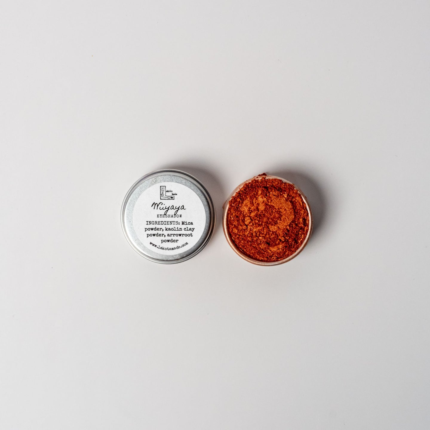 An open aluminum tin of vibrant copper orange powder on a white background with a white label that says Lakota Made Wiyaya Eyeshadow Ingredients mica powder, kaolin clay powder, arrowroot powder.