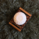 A close up of a round flower shaped white colored soap bar on a brown wooden soap dish on pine needles.