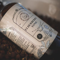 A large brown bottle with a white label that says Lakota Made Elderberry Tonic. There is a bundle of dried berries next to the bottle. It is resting on its side.