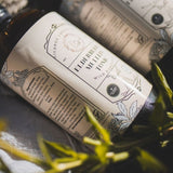 A large brown bottle with a white label that says Lakota Made Elderberry Mullein Tonic. There is a bundle of flowers next to the bottle. It is resting on it's side on a cream linen cloth.