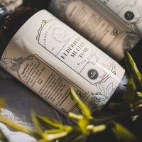 A large brown bottle with a white label that says Lakota Made Elderberry Mullein Tonic. There is a bundle of flowers next to the bottle. It is resting on it's side on a cream linen cloth.