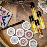 An image of a variety of makeup products on a table top with dried sage, native beadwork, abalone shell, and a makeup brush. The makeup items include round metal tins of eyeshadow and rollerball bottles of lip oil