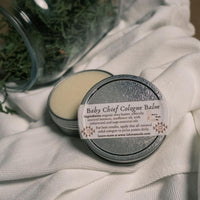 A metal tin filled with beeswax solid cologne with a white label that says Baby Chief Cologne Balm next to a glass far filled with dried leaves
