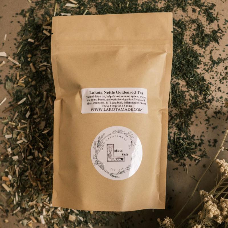 A brown bag of loose leaf tea with a white label that says Lakota Nettle Goldenrod Tea surrounded by dried flowers and herbs