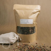 A brown bag full of dried flowers and herbs with a white label that says Rejuvenating Bath Tea next to a cotton drawstring tea bag