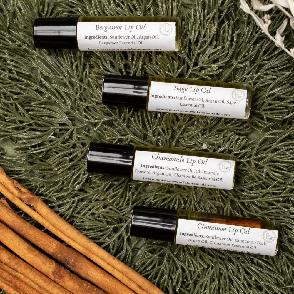 Close up image of four roller ball bottles of Lakota Made's War Paint Lip Oils. Bergamot Lip Oil, Sage Lip Oil, Chamomile Lip Oil, and Cinnamon Lip Oil. Bottles are resting on pine needles next to cinnamon sticks.