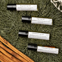 Close up image of four roller ball bottles of Lakota Made's War Paint Lip Oils. Bergamot Lip Oil, Sage Lip Oil, Chamomile Lip Oil, and Cinnamon Lip Oil. Bottles are resting on pine needles next to cinnamon sticks.