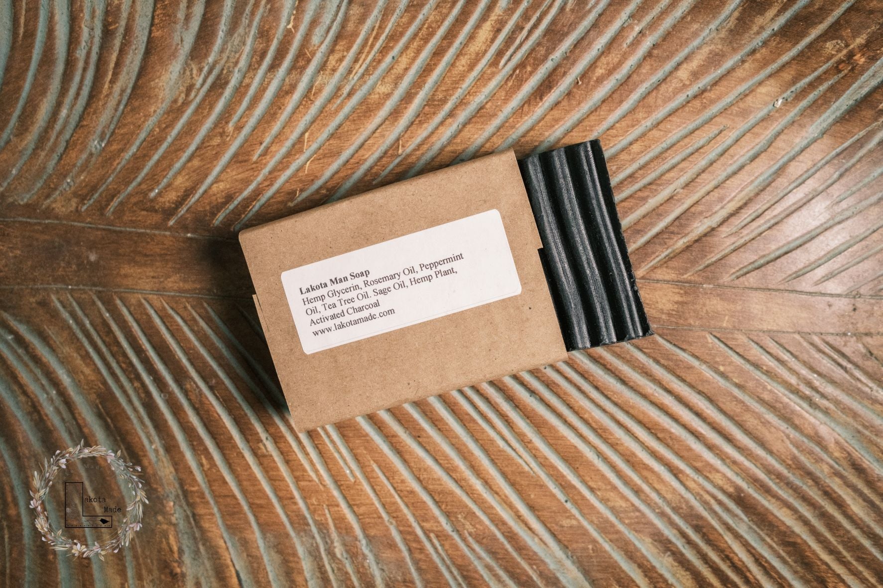 A rectangular grooved black colored soap bar with a second bar 
 in a brown paper sleeve. The package says Lakota Made Lakota Man Soap. The products are on a cloth in a wooden dish surrounded brown carved wooden palm leaf.